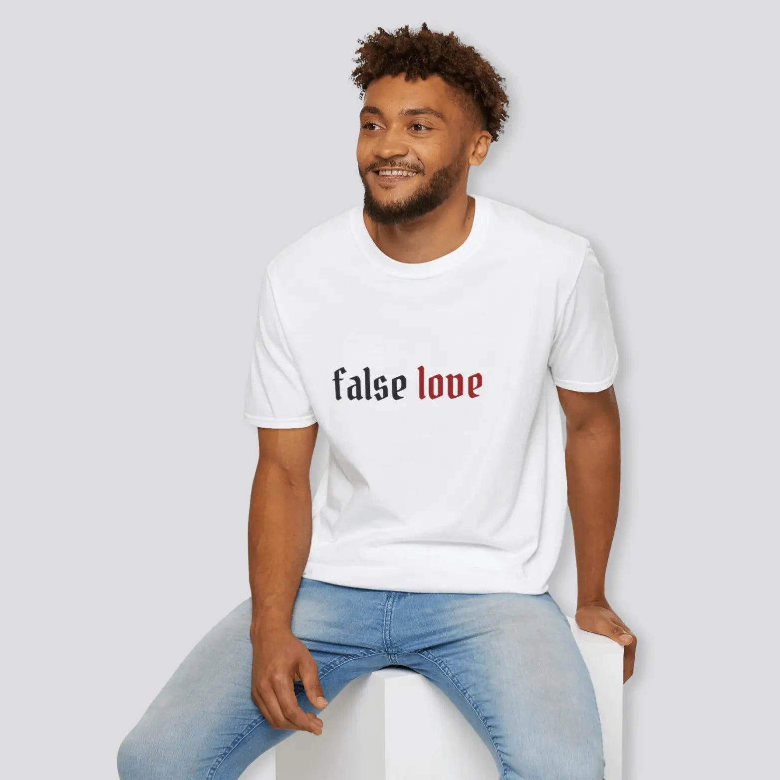 Man wearing a white t-shirt with 'false love' text, sitting on a white stool against a light gray background.
